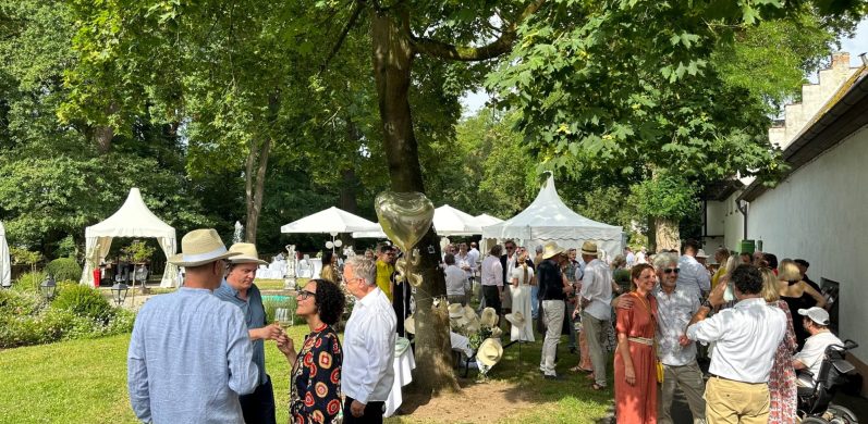 Menschen in festlicher Kleidung stehen in einem Garten mit Zelten und Bäumen.